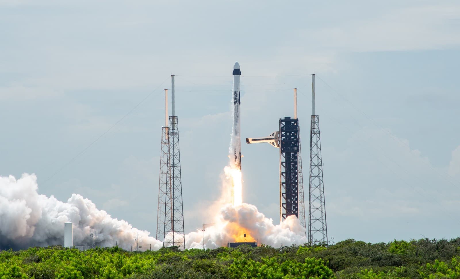 A Falcon 9 rises above its launch tower through white exhaust clouds under a pale blue sky.