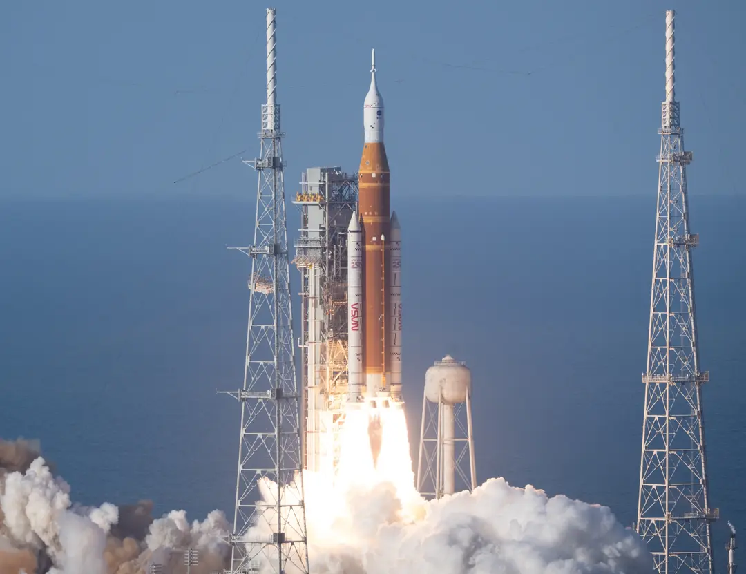 The Space Launch System rocket lifts off between launch towers, with bright exhaust below it.
