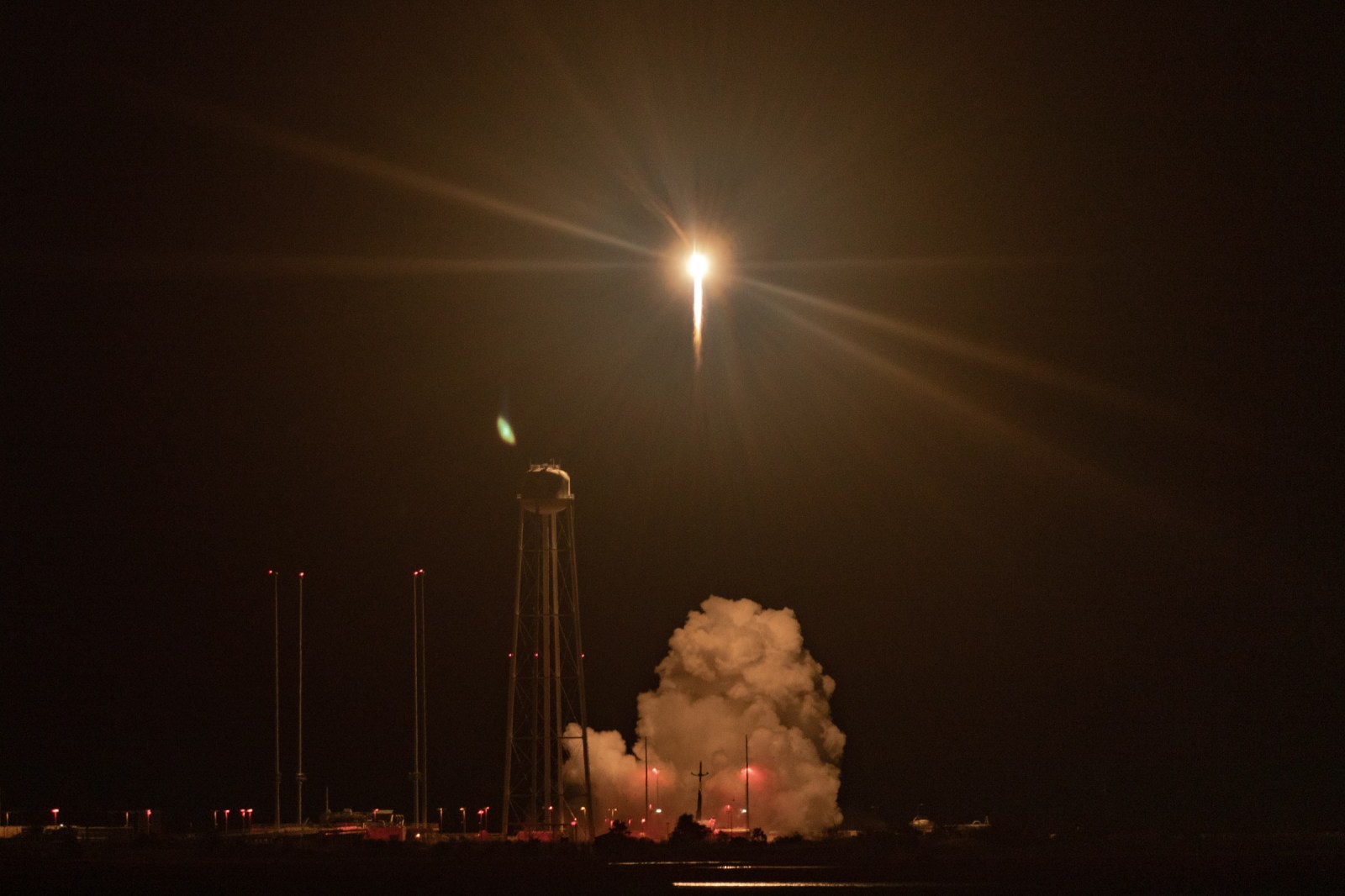 The light of an Electron launch rises above a water tower and an illuminated exhaust cloud at night.