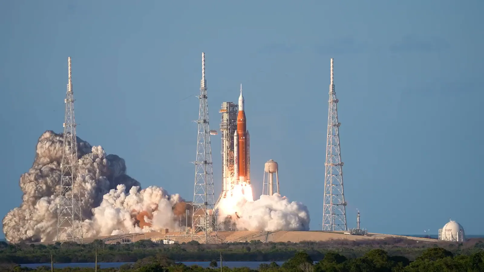 The orange and white SLS rocket rises past its launch tower on a column of white fire while exhaust clouds billow across the pad.