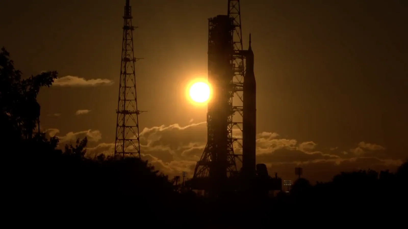 Time-lapse: the rising Sun flares behind the silhouetted rocket and its launch tower above a line of trees and golden cloud.