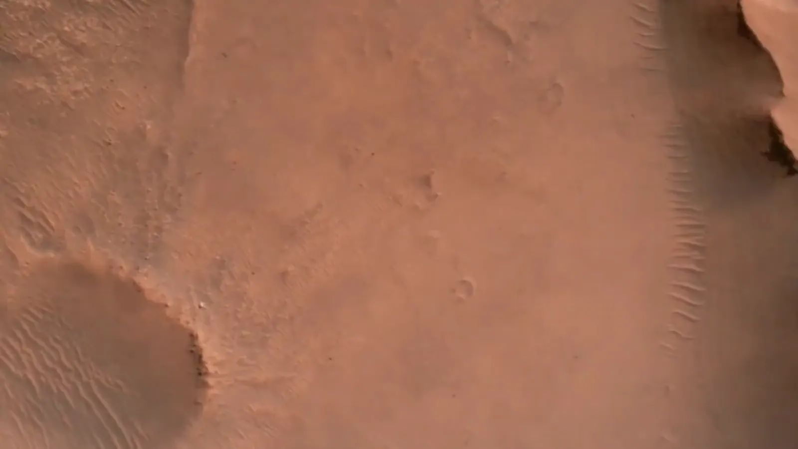 Video: seen from above, a rust-coloured plain slides past with a small crater, a pale rippled cliff edge and then a larger crater filled with ripples.