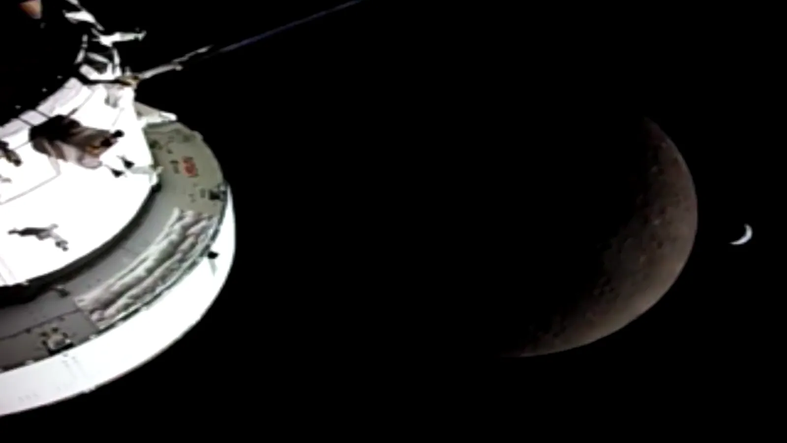 Video: the bright white hull of the Orion spacecraft at left, a dim crescent Moon at right and a tiny bright crescent Earth beside it.